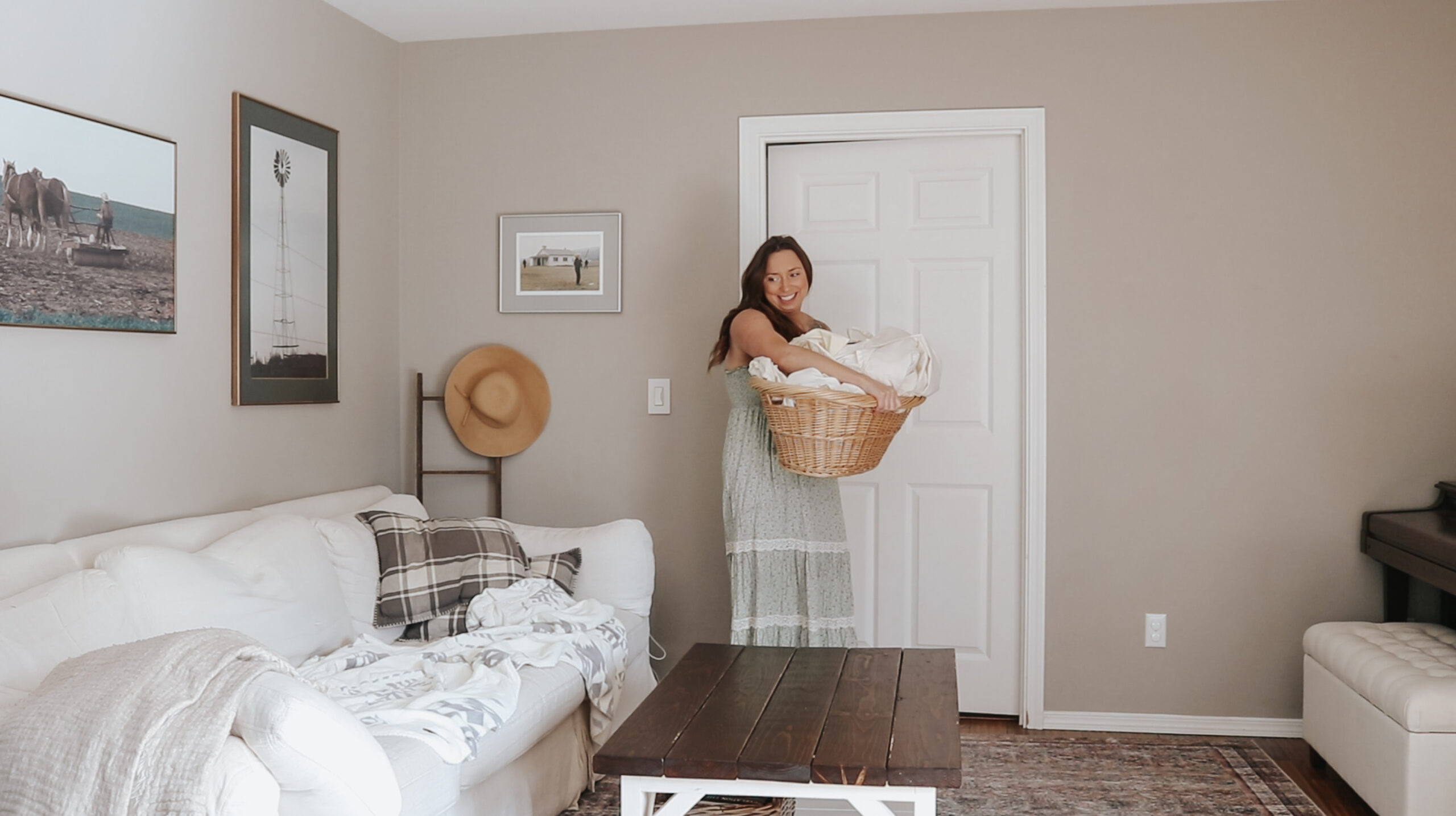 Christian homemaker carries a basket of laundry from one room to the next. She is in her living room with a peaceful environment. The couch is white and there is a coffee table in front of it.